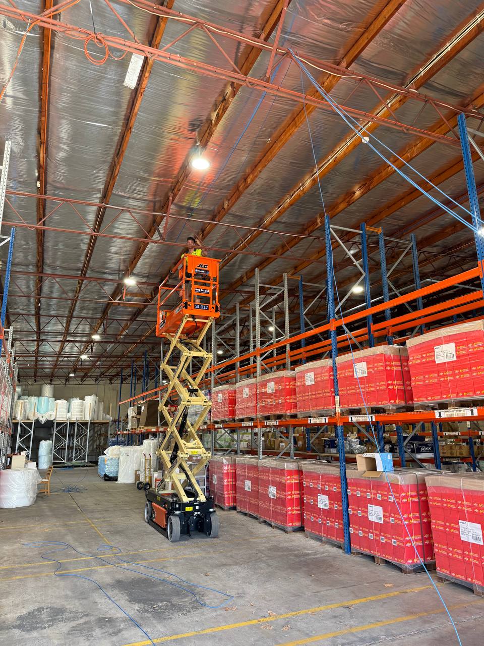 Electrician on a scissor lift working on high-bay lighting in a warehouse, with pallet racking below