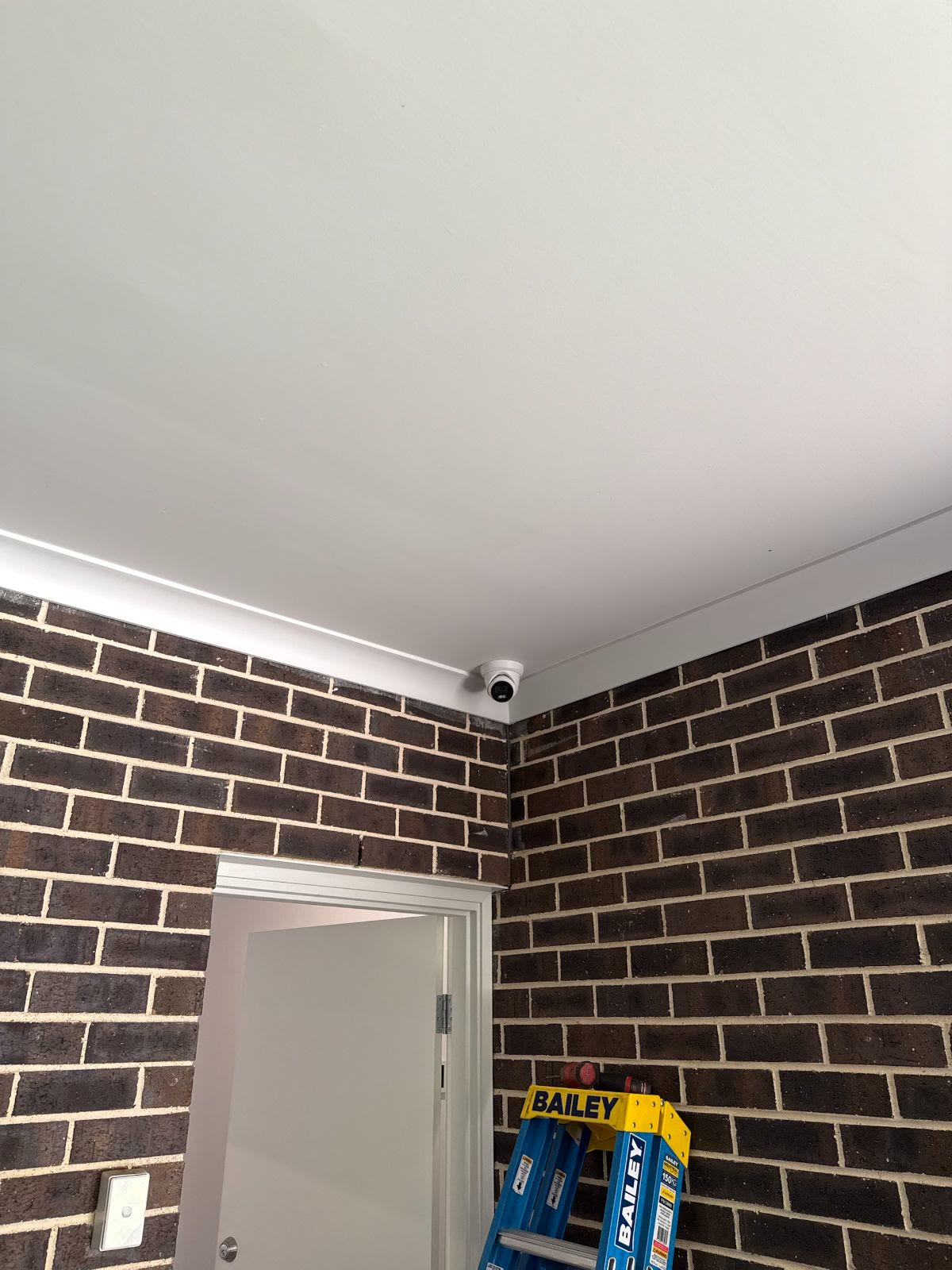 Turret camera mounted in the ceiling corner of a brick-walled room, aimed at the doorway